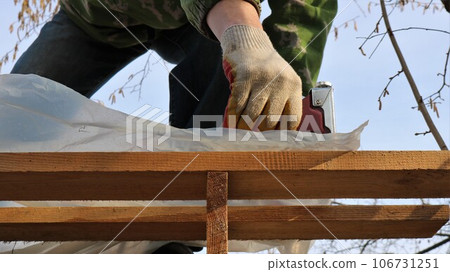 fixing plastic film on the wooden floors of the roof of a barn frame using a construction stapler, a construction gun in the gloved hands of a builder in the process of fastening polyethylene fixing plastic film on the wooden floors of the roof of a barn frame using a construction stapler, a construction gun in the gloved hands of a builder in the process of fastening polyethylene 106731251