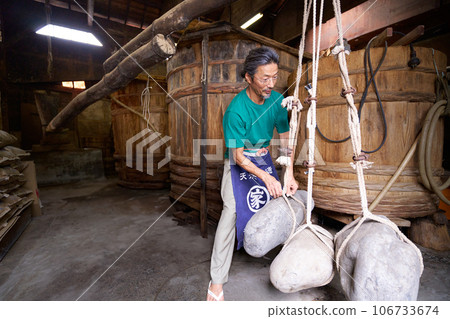 工匠用石鎚從醪中榨出醬油。傳統製造方法啤酒廠的圖片。 工匠用石鎚從醪中榨出醬油。傳統製造方法啤酒廠的圖片。 106733674