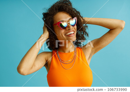 Positive woman in heart-shaped glasses touching curly hair standing on blue studio background Positive woman in heart-shaped glasses touching curly hair standing on blue studio background 106733990