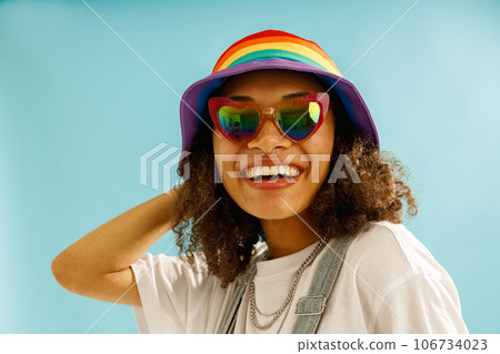 Smiling woman in rainbow cap and heart-shaped glasses looks camera over blue studio background Smiling woman in rainbow cap and heart-shaped glasses looks camera over blue studio background 106734023