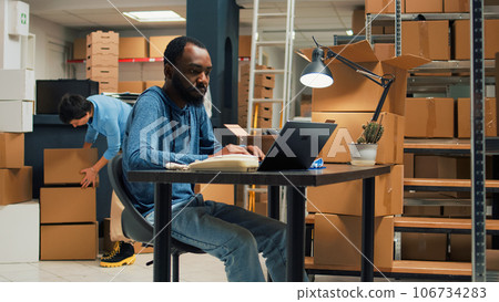 African american worker planning logistics to ship products, preparing order shipment with stock inventory information. Male owner working on laptop with packages from warehouse racks. African american worker planning logistics to ship products, preparing order shipment with stock inventory information. Male owner working on laptop with packages from warehouse racks. 106734283