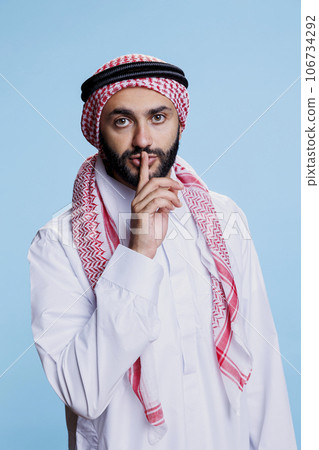 Muslim man wearing traditional clothes showing quiet gesture and touching finger to lips portrait. Arab person making tsss sign and looking at camera with serious expression 106734292