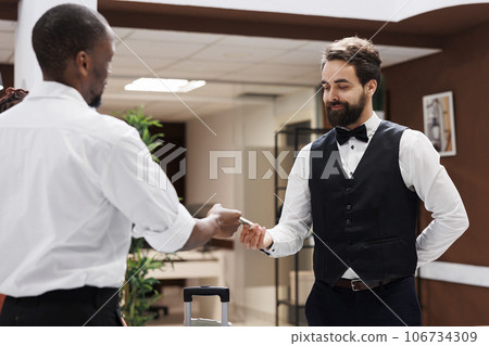 Hotel porter taking money from client, accepting to carry luggage and help with bags after check in process. Male steward working as bellboy at luxury hotel, receiving cash tip from guests. Hotel porter taking money from client, accepting to carry luggage and help with bags after check in process. Male steward working as bellboy at luxury hotel, receiving cash tip from guests. 106734309
