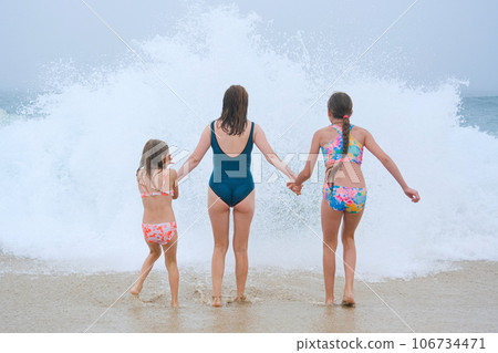 Mother and children playing on the ocean beach. Family enjoying the ocean. Mother holds girls's hands and they all look at the ocean together. 106734471