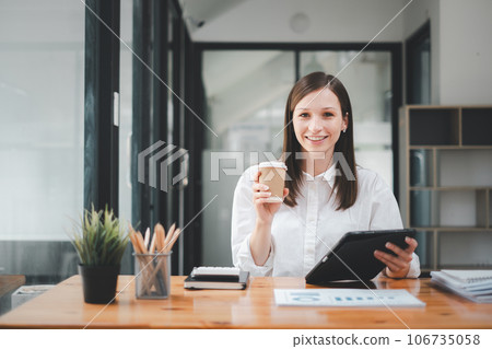 Smiling confident business leader looking at camera and sittingin an office, Portrait of confident businesswoman hand holding coffee cup in boardroom. Using digital tablet during a meeting. Smiling confident business leader looking at camera and sittingin an office, Portrait of confident businesswoman hand holding coffee cup in boardroom. Using digital tablet during a meeting. 106735058