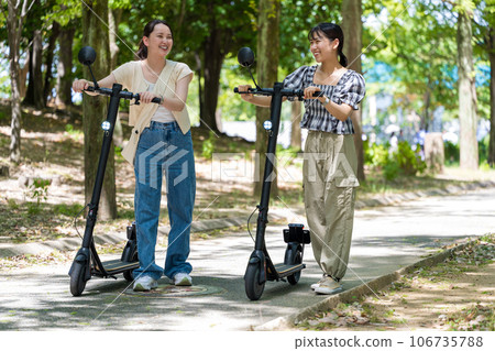 Two young and cute women traveling on electric kickboards along a tree-lined avenue with beautiful greenery | Electric kickboard image 106735788