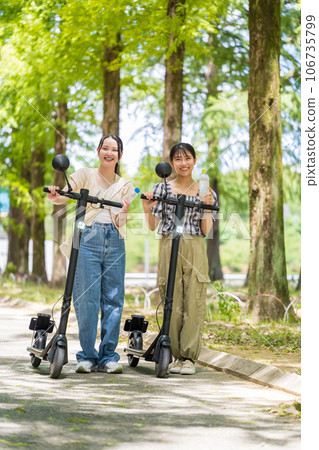 Two cute women taking a walk on a beautiful green tree-lined street on electric kickboards and rehydrating themselves | Electric kickboard image Two cute women taking a walk on a beautiful green tree-lined street on electric kickboards and rehydrating themselves | Electric kickboard image 106735799