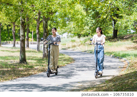 Two young and cute women running on electric kickboards along a tree-lined avenue with beautiful greenery | Electric kickboard image Two young and cute women running on electric kickboards along a tree-lined avenue with beautiful greenery | Electric kickboard image 106735810