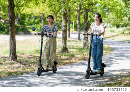 Two young and cute women running on electric kickboards along a tree-lined avenue with beautiful greenery | Electric kickboard image Two young and cute women running on electric kickboards along a tree-lined avenue with beautiful greenery | Electric kickboard image 106735812