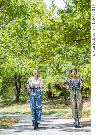 Two young and cute women running on electric kickboards along a tree-lined avenue with beautiful greenery | Electric kickboard image 106735817