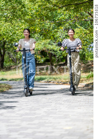 Two young and cute women running on electric kickboards along a tree-lined avenue with beautiful greenery | Electric kickboard image 106735818