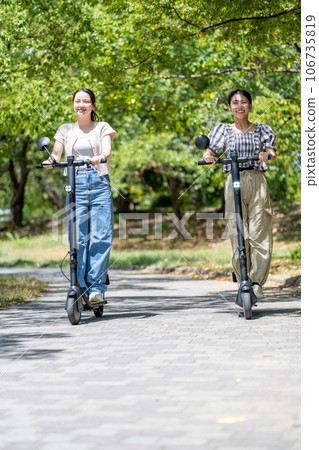 兩位年輕可愛的女士在綠樹成蔭的綠樹成蔭的大道上騎著電動踢水板跑步 | 電動踢水板圖片 106735819