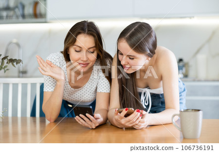Happy mother and teen daughter looking at their phones leaning on kitchen table 106736191