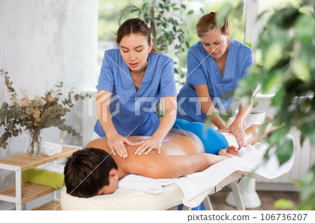 Young masseuse in tandem with female colleague performing back massage to man 106736207