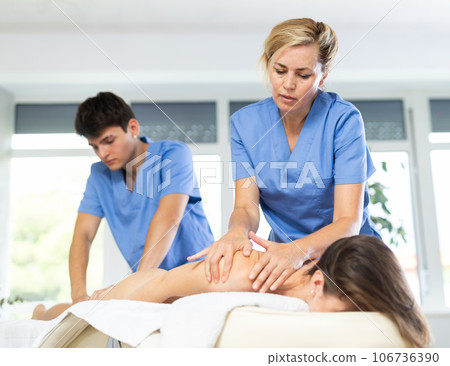 Two female and male masseuses perform stroking and rubbing of muscles of back and shoulders to girl Two female and male masseuses perform stroking and rubbing of muscles of back and shoulders to girl 106736390