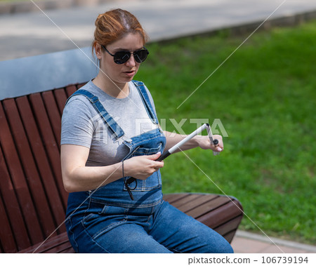 Blind pregnant woman folding walking stick while sitting on bench. Blind pregnant woman folding walking stick while sitting on bench. 106739194