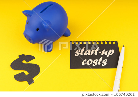 Business concept. On a yellow background there is a piggy bank, next to it is a dollar symbol and a leaf from a notepad with the inscription - start-up costs Business concept. On a yellow background there is a piggy bank, next to it is a dollar symbol and a leaf from a notepad with the inscription - start-up costs 106740201