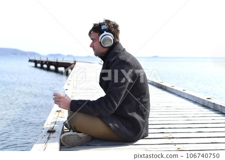 Young man listening to music and drinking coffee, sitting alone on pier on a sunny day 106740750