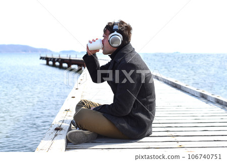 Young man listening to music and drinking coffee, sitting alone on pier on a sunny day Young man listening to music and drinking coffee, sitting alone on pier on a sunny day 106740751