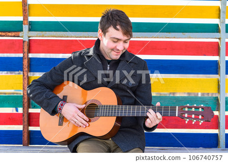 Smiling young man playing guitar and sitting against colorful wall 106740757