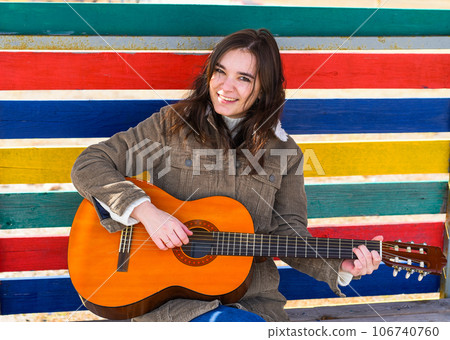 Young woman playing guitar and laughing, sitting on a bench against colorful background 106740760
