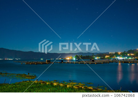 The evening atmosphere at Lake Batur, Kintamani, Bali, with the reflection of Mount Batur and the ambiance of the village along the lake's edge, illuminated by sparkling lights. 106742868