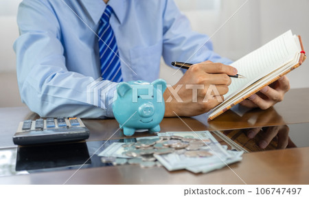 Businessman hand writing income and expenses to account with piggy bank and dollar notes, step up start-up businesses to success, Saving money for future plan and retirement fund concept Businessman hand writing income and expenses to account with piggy bank and dollar notes, step up start-up businesses to success, Saving money for future plan and retirement fund concept 106747497
