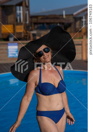An elderly woman in a huge straw hat and sunglasses is sunbathing by the pool. Retiree on vacation. An elderly woman in a huge straw hat and sunglasses is sunbathing by the pool. Retiree on vacation. 106749183