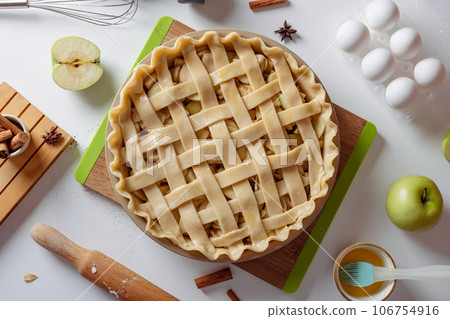 Uncooked apple pie into a baking dish ready for cooking on a white table surrounded by ingredients 106754916
