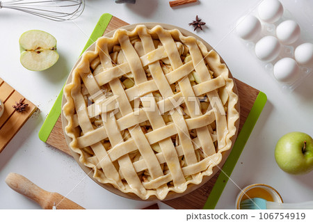Uncooked apple pie into a baking dish ready for cooking on a white table surrounded by ingredients 106754919