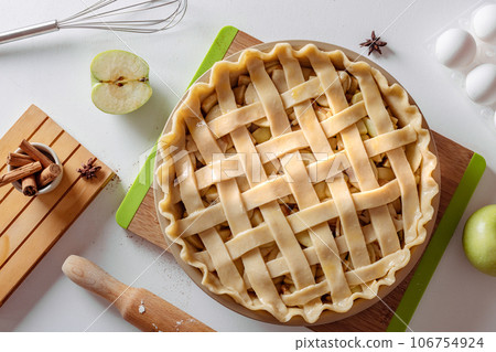 Uncooked apple pie into a baking dish ready for cooking on a white table surrounded by ingredients 106754924