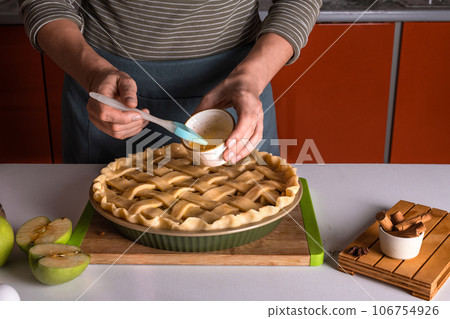 Woman spreading eggs on the raw apple pie with a cooking brush in a bakery dish, recipe 106754926
