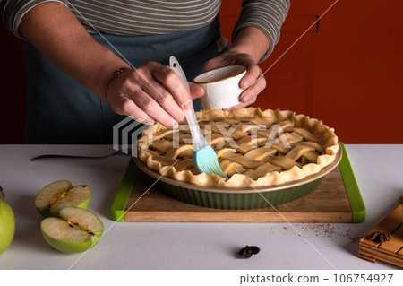 Woman spreading eggs on the raw apple pie with a cooking brush in a bakery dish, recipe 106754927