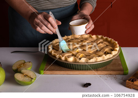 Woman spreading eggs on the raw apple pie with a cooking brush in a bakery dish, recipe 106754928