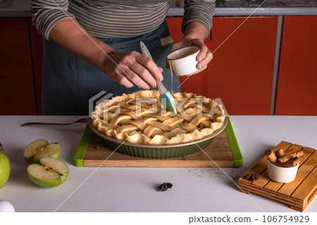 Woman spreading eggs on the raw apple pie with a cooking brush in a bakery dish, recipe 106754929