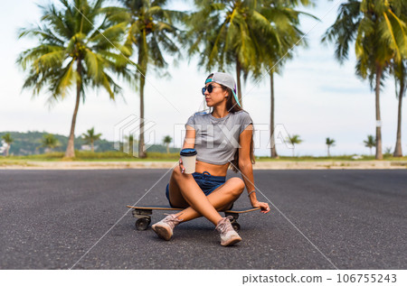 Young beautiful woman in a cap sitting on a skateboard and drinking coffee Young beautiful woman in a cap sitting on a skateboard and drinking coffee 106755243