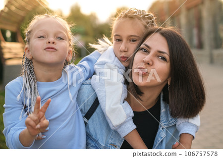 Two cute little preschool girls hug their mother and blow a kiss. Loving mom on a walk with children Two cute little preschool girls hug their mother and blow a kiss. Loving mom on a walk with children 106757578
