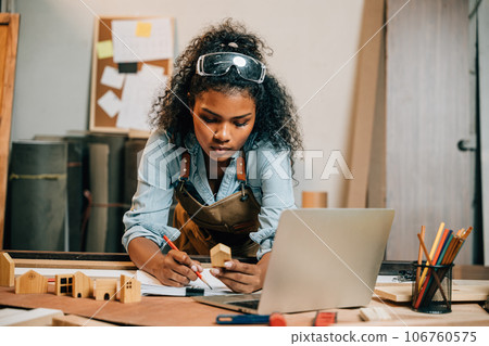 Carpenter america black woman curly hair sketch making notes in work paper while standing at desk with laptop computer in wood workshop, young female working learning online at woodshop 106760575