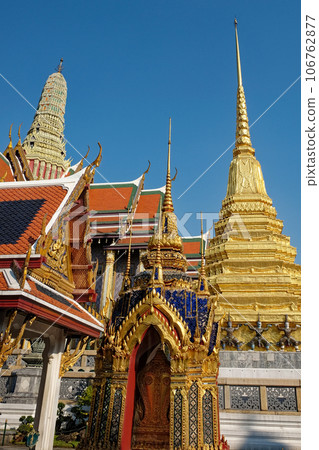 A picturesque low-angle view offers a glimpse of a Thai temple's distinctive red roofs, intricate gold accents, and a towering white spire standing tall beneath a serene blue sky. 106762877