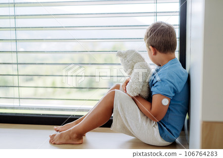 Diabetic boy with a continuous glucose monitor sitting by the window, holding his stuffed teddy bear and looking outside. Diabetic boy with a continuous glucose monitor sitting by the window, holding his stuffed teddy bear and looking outside. 106764283