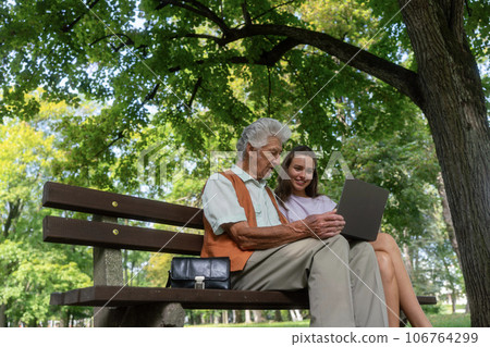 The grandfather and granddaughter are looking at photos on notebook, sitting on a bench in a city park. 106764299