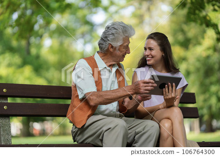 The grandfather and granddaughter are looking at photos on a tablet, sitting on a bench in a city park. 106764300