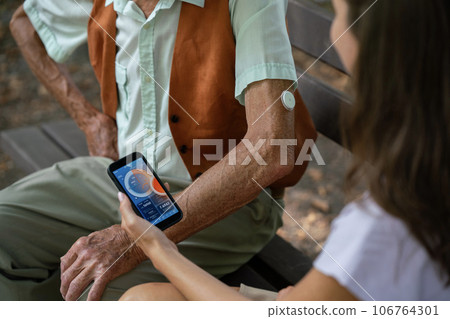 Caregiver helping senior diabetic man check his glucose data on smartphone outdoors, in park. 106764301