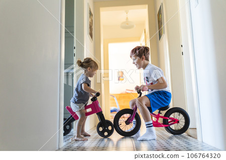 Siblings are riding bicycles indoors, in the hallway of the house. 106764320