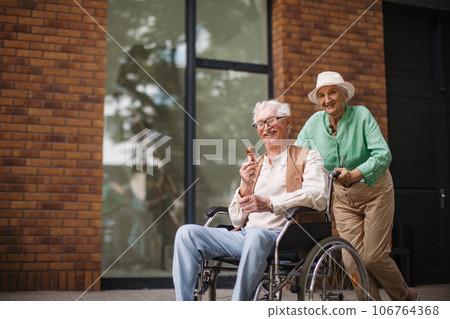 Elderly woman pushing her delightful husband in wheelchair. 106764368