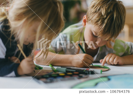 Siblings lying on stomach and painting at home with watercolors, markers and tempera paints. 106764376