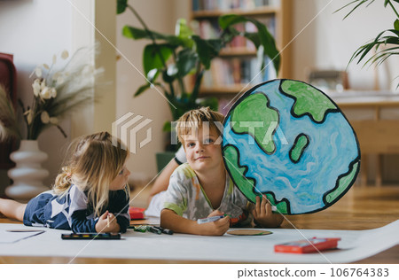 Siblings lying on stomach and painting at home with watercolors, markers and tempera paints, creating a model of planet Earth. 106764383