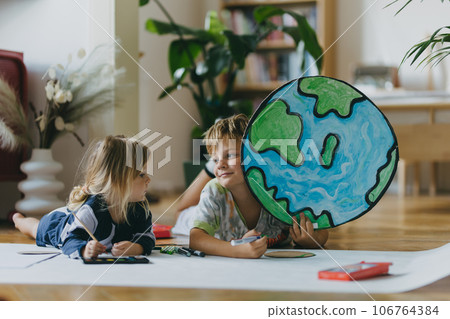 Siblings lying on stomach and painting at home with watercolors and tempera paints, creating a model of planet Earth. 106764384