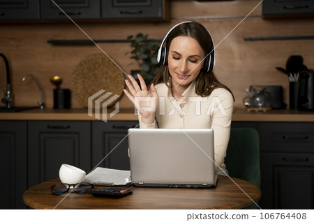 A young brunette woman wearing headphones talks via a laptop via video link while sitting in the kitchen A young brunette woman wearing headphones talks via a laptop via video link while sitting in the kitchen 106764408