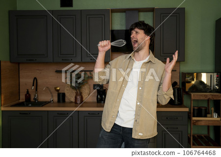 A funny guy sings a song, holding a mixer with a microphone in his hands, listening to music in a modern kitchen, happy joyful young people are having fun cooking A funny guy sings a song, holding a mixer with a microphone in his hands, listening to music in a modern kitchen, happy joyful young people are having fun cooking 106764468